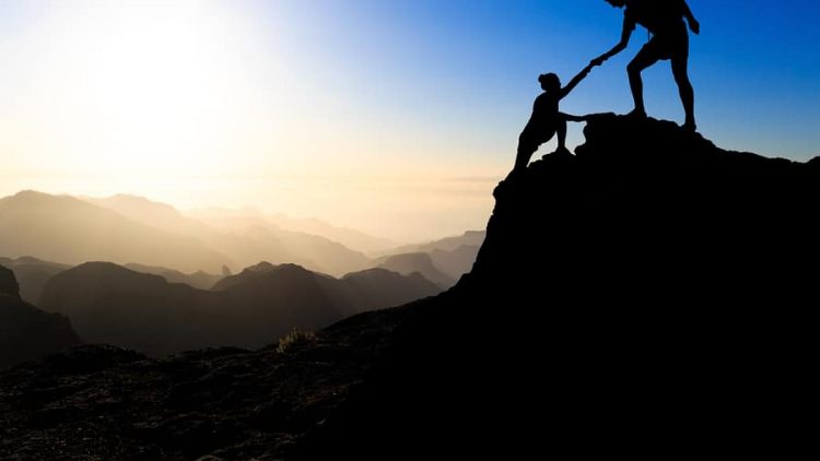 a man and a woman hiking up a mountain during a sunset where the man is helpign the woman up. this image is being used to convey the importance of working together during collaborative divorce to achieve win-win outcomes during the divorce.